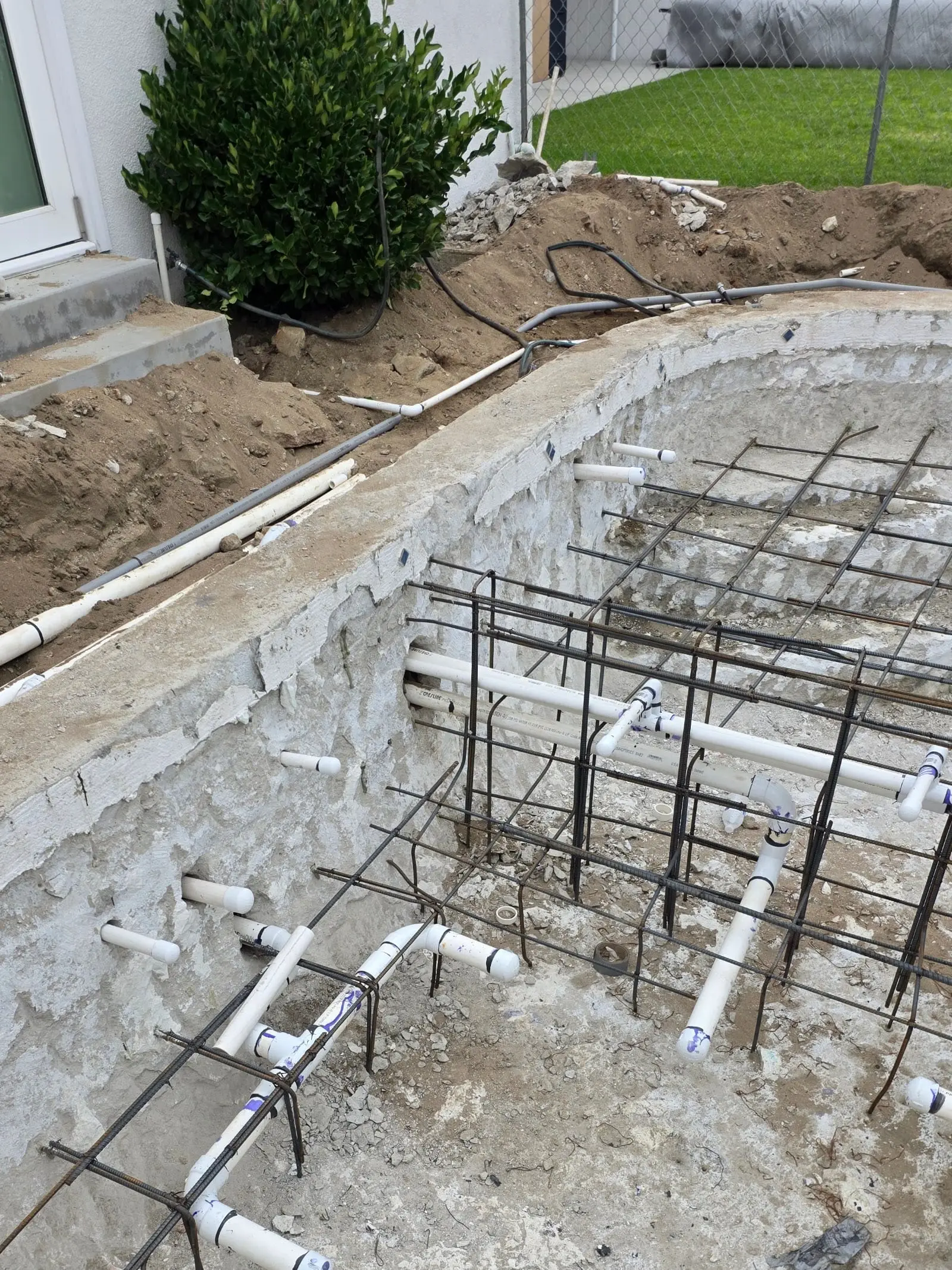 Excavation with a rough concrete wall, white PVC pipes laid through steel rebar, capped pipe stubs set into the wall and trenches with pipe and flexible conduit beside a house