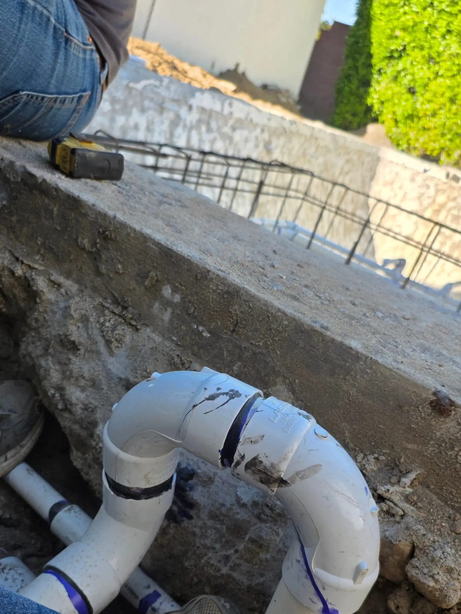 White PVC elbows and pipe joined together in an excavation below a concrete edge, with steel rebar beyond and a person in jeans and sneakers at the edge of the frame