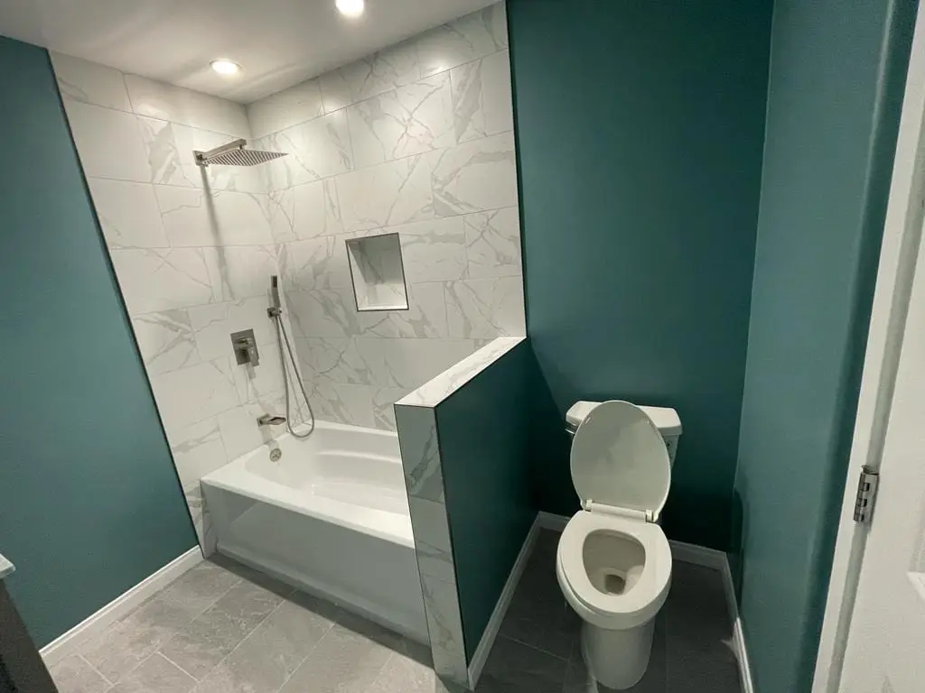 Bathroom with teal walls, a bathtub with a white marble-look tiled surround, a square rain shower head and a handheld sprayer, a tiled half wall and a toilet