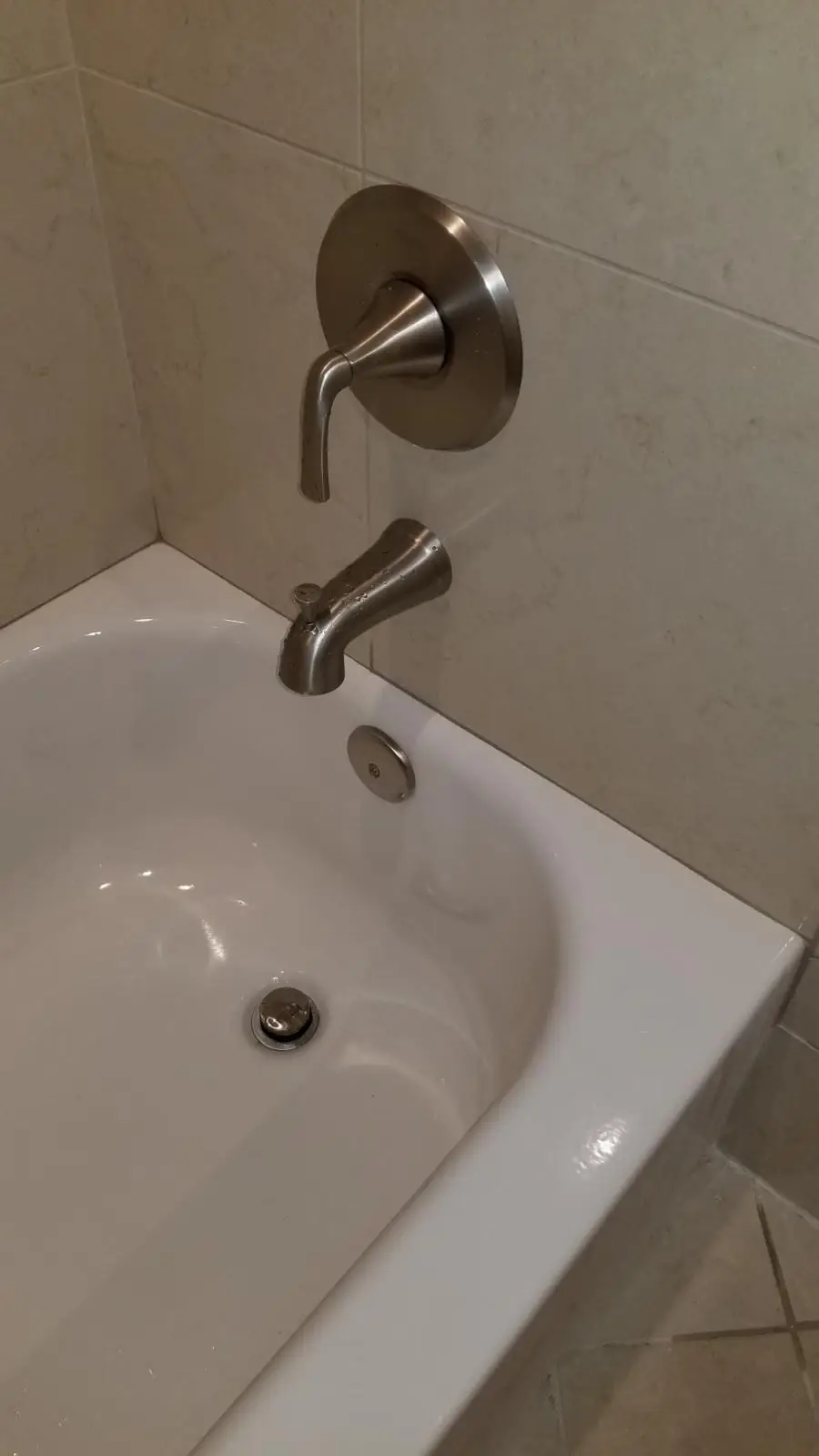 Brushed nickel tub spout and single lever valve on a beige tiled wall above the corner of a white bathtub with its drain