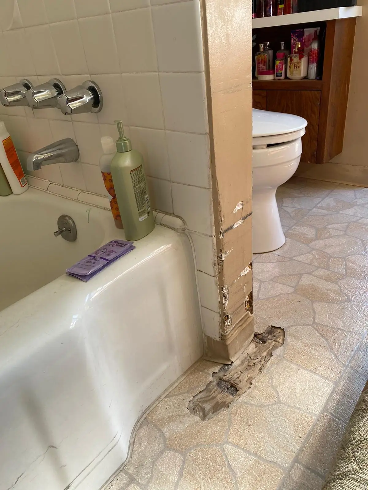 Corner of a bathroom wall beside a bathtub with chipped paint and broken, discolored wood at its base where it meets the vinyl floor